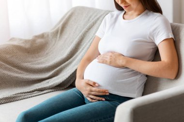 Doğum Konsepti. Genç, güzel bir kadın bebek bekliyor, evinde koltukta dinleniyor, mutlu, gülümseyen bir kadın hamile karnını şefkatle kucaklıyor, rahat bir koltukta dinleniyor, boşluğu kopyalıyor.