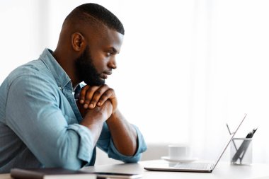 İçişleri Bakanlığı 'nda laptopla çalışan düşünceli bir siyah yazar. Afrika kökenli Amerikalı serbest çalışan adam masada oturuyor ve ekran ekranına bakıyor. Pahalı bir yüz ifadesi, kopyalama alanı ile yan görünüm.