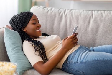 Mutlu siyah bayan kanepede cep telefonuyla uzanıyor, arkadaşlarıyla sohbet ediyor, patlamış mısır ve fincanla kahve ya da çay içiyor, oturma odası içi, yan görüş, kopyalama alanı.