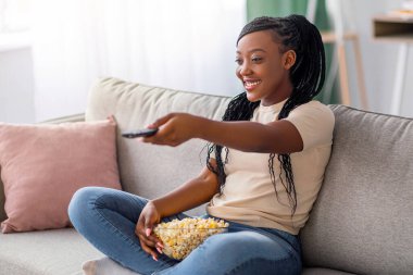 TV kumandalı koltukta oturan mutlu genç siyahi kadın, izlemek ve yemek için film seçiyor, patlamış mısır yiyor, fotokopi çekiyor. Neşeli Afrikalı Amerikalı kadın kanal değiştiriyor, ev eğlencesi konsepti