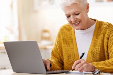 Çevrimiçi eğitim, kurslar, internet konsepti. Mutfakta dizüstü bilgisayarın önünde oturan, notlar alan, gülümseyen, çay içen, internetten ders çalışmaktan zevk alan yaşlı bir kadın.