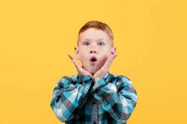 Gündelik gömlekli, yüz ifadesi olan küçük bir çocuk. Şok olmuş kızıl saçlı çocuk ağzını açık tutuyor ve yüzüne dokunuyor, şok edici bir şey görüyor, sarı stüdyo arka planı, kopyalama alanı..