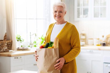 Gülümseyen yaşlı kadın COVID-19 salgını sırasında internetten sipariş edilen poşetleri kontrol ediyor. Mutfağın içi, fotokopi alanı. Alışverişten sonra paketleri açan neşeli yaşlı bayan. Yemek teslimatı