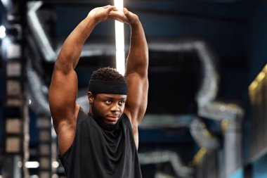 Kas yığını Afrikalı Amerikalı genç vücut geliştirici beden eğitimi yapıyor, kollarını uzatıyor ve fotokopi odasına bakıyor. Yakışıklı zenci sporcu antrenmandan önce hazırlanıyor, ısınıyor.