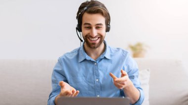 Video çağrısı. Kulaklıklı mutlu adam içeride oturan dizüstü bilgisayarla konuşuyor. Uzaklık Çalışması ve E-Öğrenme, Çevrimiçi Eğitim ve İş Eğitimi Konsepti. Seçici odak
