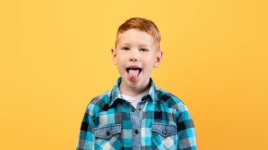 Şımarık kızıl çocuk, günlük kıyafetli tatlı okul çocuğu sarı stüdyo arka planında dilini kameraya gösteriyor, kopyalama alanı. Şımarık çocuk, çocukların davranış sorunları