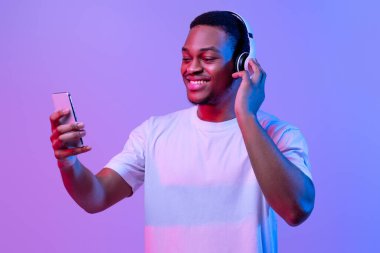 Müzik Yayını Uygulaması. Kablosuz Kulaklıklı Yakışıklı Siyah Adam Smartphone 'da Favori Şarkıları Dinliyor, Çevrimiçi Mobil Uygulama Kullanıyor, Mor Arkaplan Üzerindeki Neon Işıklandırması Altında Duruyor, Serbest Alan