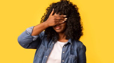 Kıvırcık siyah kadın tek eliyle gözlerini kapatıyor, korkmuş ya da endişeli hissediyor, merak ediyor ya da körü körüne sarı stüdyo arkaplanına karşı bir sürpriz bekliyor.