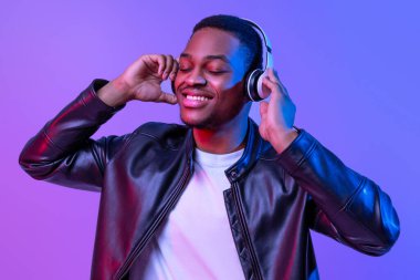Müzik aşığı. Kablosuz kulaklıklı yakışıklı siyah adam ve en sevdiği şarkıları dinleyen deri ceketli rahat Afro-Amerikan adam, Mor Arkaplan Üzerindeki Neon Işığı Altındaki Kapalı Gözler