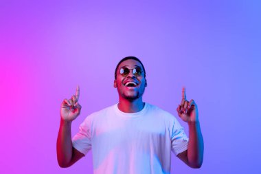 Neon ışıklarının altında dikilen heyecanlı siyahi adam fotokopi uzayını işaret eden, şık güneş gözlüklü neşeli Afrikalı Amerikalı adam, Mor Arkaplan üzerinde boş yer gösteriyor