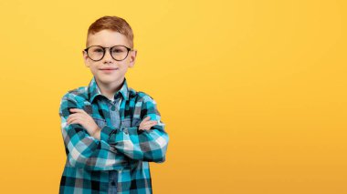 Kolları göğsünde çapraz duran, sarı stüdyo arka planında poz veren, gözlüklü, zeki kızıl çocuk. Sevimli, kızıl saçlı, ekose gömlekli inek çocuk kameraya gülümsüyor, boşluğu kopyalıyor.