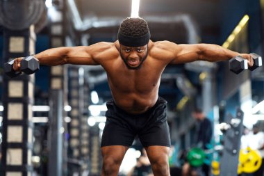 Güçlü üstsüz zenci vücut geliştirici spor salonunda dambıllarla egzersiz yapıyor. Duygusal Afrikalı Amerikalı atletik adam halterleri kaldırıyor, kol ve sırt kasları üzerinde çalışıyor, boşluğu kopyalıyor.