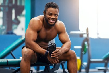 Gülümseyen Afro-Amerikan üstsüz vücut geliştirici modern spor salonunda çalışırken protein ya da su içerek. Egzersiz sonrası dinlenen mutlu siyah adam, kopyalama alanı