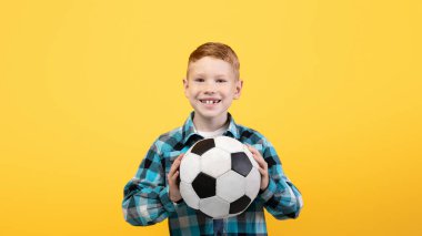 Genç futbolcu. Sarı arka planda futbol topuyla poz veren neşeli çocuk, fotokopi alanı. Futbol topunu kameraya gösteren mutlu çocuk, çocuklar ve gençler spor yaşam tarzı konsepti