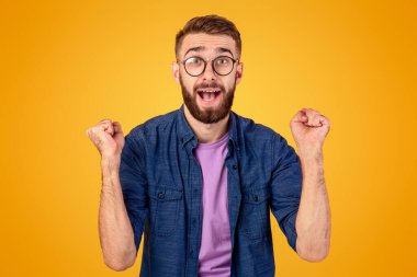 Kazanan ve başarı konsepti. Heyecanlı genç adam, turuncu stüdyo arka planında yumruklarını sıkarak EVET hareketi yapıyor. Heyecanlı Kafkasyalı adam büyük bir zaferle sevinçle bağırıyor.