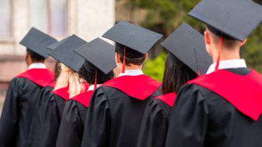 Mezuniyet cübbesi ve şapkalı uluslararası öğrenci grubunun üniversite kampüsünün önünden geçişi, yakın plan. Mezuniyet törenine katılan tanınmamış çok ırklı öğrenciler