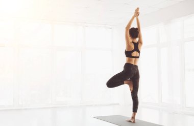 Bir kadın, parlak, minimalist ve geniş pencereli bir stüdyoda sabah yoga seansı sırasında ağaç pozu vererek doğal ışığın boşluğu doldurmasına, arka plana bakmasına, boşluğu kopyalamasına izin verir.