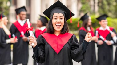 Mezuniyet kostümü giymiş mutlu Asyalı genç bayan diplomasını gösteriyor ve kameraya bağırıyor. Üniversite kampüsünde uluslararası bir grup öğrenciye poz veren mutlu bayan öğrenci.