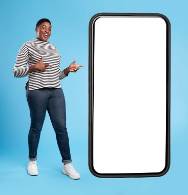 Neşeli Afrikalı Afro-Amerikan Kadın Büyük Akıllı Telefonun yanında boş ekranlı ve parmakla işaret eden Mobil Uygulama Mavi Stüdyo Arkaplanının Üzerinde Duruyor. Kare çekim, Mockup