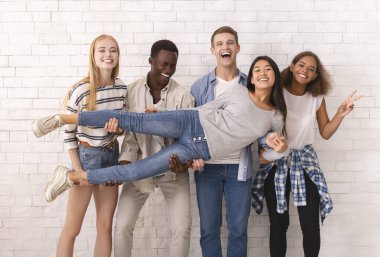 Duygusal uluslararası öğrenci grubu beyaz tuğla duvar arkasında eğleniyor, Asyalı kızı kaldırıyor.