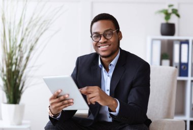 Gülümseyen siyah takım elbiseli genç adam dijital tablet tutuyor, modern teknolojileri iş dünyasında, ofis içinde, kopyalama alanında kullanıyor. Neşeli Afro-Amerikan iş adamı iş için uygulama kullanıyor.