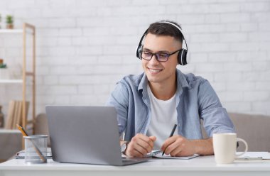 Kişisel izolasyon sırasında evde yeni bilgiler. Gözlüklü ve kulaklıklı mutlu genç adam notlar alıyor ve oturma odasındaki dizüstüne bakıyor.