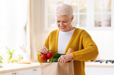 Gülümseyen yaşlı bayan COVID-19 karantinası, mutfak içi, fotokopi alanı sırasında internetten sipariş edilen poşetleri kontrol ediyor. Paketleri açan mutlu yaşlı kadın. Yiyecek teslimatı kavramı
