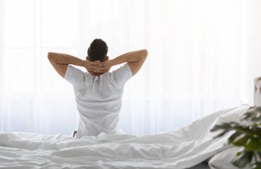 İyi sabahlar. Uyandıktan sonra elleri başının arkasında oturan, beyaz tişörtlü, aydınlık yatak odasında esneyen, tanınmayan bir erkek görüntüsü, Boşluğu Kopyala