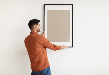 Modern Ev İçi ve İç Dekorasyon. Gülümseyen genç adam resim asıyor, duvara resim çerçevesi asıyor. Boş poster, tasarım modeli, boş kopya alanı gösteren sıradan bir adam.