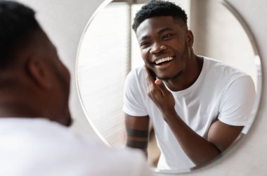 Evdeki modern banyoda dikilirken aynadaki yansımasına gülümseyerek tıraş olmadan önce tıraşsız Afrikalı Amerikalı adamın çenesine tüy değdirmesi. Erkek Cilt Bakımı. Seçici Odaklanma