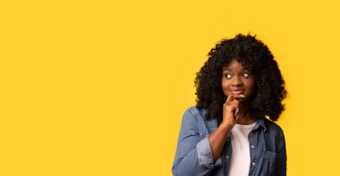 Gayri resmi kıyafetli, fotokopi odasına bakan, çenesine dokunan, sarı stüdyo arkaplanı, panorama olan güzel siyahi bir kadın. Güzel Afro-Amerikan kıvırcık bayan reklamcılık için heyecanlı.