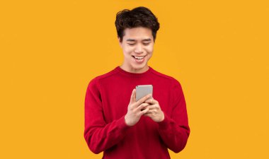 Halk ve Teknoloji Konsepti. Portakal rengi stüdyo arka planında izole edilmiş akıllı telefon kullanan mutlu Asyalı genç adam portresi. İnternette sohbet eden, sosyal medyada gezinen