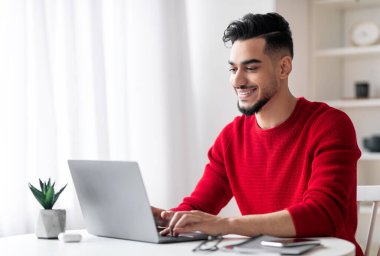 İçişleri Bakanlığı 'nda dizüstü bilgisayarda çalışan yakışıklı Arap erkek serbest çalışan, bilgisayarla aydınlık odada oturan ve klavyede yazan, uzaktan çalışmanın keyfini çıkaran, boşluğu kopyalayan gülümseyen orta doğulu genç adam.