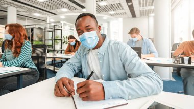 Yeni kurallar, önlemler. Afro-Amerikan erkek öğrencinin portresi masada oturuyor, deftere yazıyor ve kameraya bakıyor, tek kullanımlık tıbbi maske takıyor, afro adam notlar alıyor.
