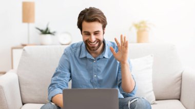 Video çağrısı. Adam el sallayarak dizüstü bilgisayara bağlanıyor ve evdeki koltukta oturuyor. Video konferansı yapan bilgisayarlara merhaba diyen adam. Modern Uzak İletişim Konsepti