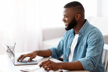 Çevrimiçi Eğitim. Genç, gülümseyen siyah adam dizüstü bilgisayarla çalışıyor ve not alıyor, Milenyum Afrikalı yakışıklı Afrikalı Amerikalı adam Webinar 'ı izliyor ve bilgisayardan bilgi alıyor,
