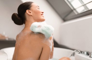 Banyo içinde dinlenen köpük köpükleriyle kaplı kadın banyosu ve yıkanma manzarası. Bayan evde banyo yapmaktan zevk alıyor. Vücut Bakımı ve Spa Konsepti. Süngere Seçici Odaklanma
