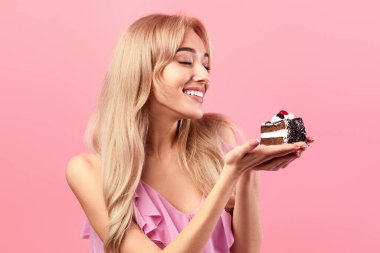 Çekici beyaz kadın elinde lezzetli bir pasta tutuyor, doğum gününde en sevdiği tatlıyı yiyor, pembe stüdyo geçmişi var. Güzel sarışın kadın özel durumu kutluyor, eğleniyor.