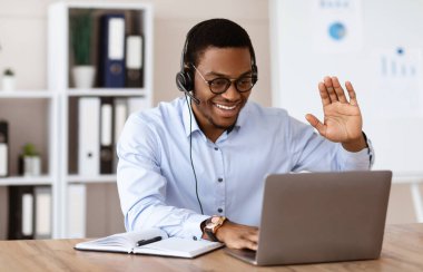 Neşeli siyahi adam iş ortaklarını selamlıyor, ofisten online konferans veriyor, dizüstü bilgisayar ve kulaklık kullanıyor, kopyalama alanı kullanıyor. Genç Afrikalı Amerikalı adam çevrimiçi eğitime katılıyor.