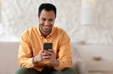 Havalı Gadget ve Uygulama. Elinde cep telefonu olan gülümseyen Arap 'ın portresi. Mesajlar yazıyor. Oturma odasındaki kanepede oturuyor. İnternette gezinen adam, internette sörf yapan, uygulamayı kullanan, boş alan