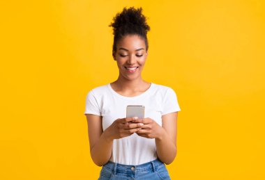 İnsanlar, Teknoloji ve İletişim Konsepti. Cep telefonuyla mesajlaşan güzel siyahi bir kadının portresi, sarı stüdyo arka planında izole edilmiş, kopyalama alanı. Sosyal medyada gülümseyen kadın sohbeti