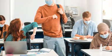 Dirsek çarpması. Muhtelif çok kültürlü insanlar, sınıfta birbirini selamlayan koruyucu yüz maskeleri takıyorlar. Sırt çantalı Asyalı çocuklar, masada oturan bir kadın, öğrenciler dokunmaktan kaçınıyor.