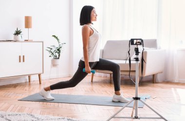 Çevrimiçi Antrenman Konsepti. Aktif Asyalı bayan kamerayla evde eğitim videosu çekiyor, dambıllarla hamleler yapıyor, yoga minderiyle egzersiz yapıyor. Oturma odasında internet kanalı için eğitim videosu kaydediliyor