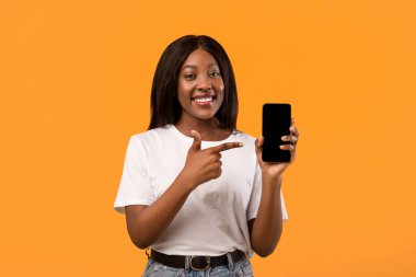 Gülümseyen genç siyahi bayan modern cep telefonunu gösteriyor, boş ekrana işaret ediyor, güzel mobil uygulamalar, turuncu stüdyo arka planı, kopyalama alanı kullandıktan sonra pozitif duygularını paylaşıyor.