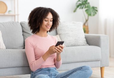 Mutlu binlerce yıllık Afro-Amerikan kadın rahat oturma odasında dinleniyor, akıllı telefonu elinde tutuyor. Genç bayan sosyal ağlarda sohbet ediyor, komik videolar izliyor, evde mobil uygulama kullanıyor.