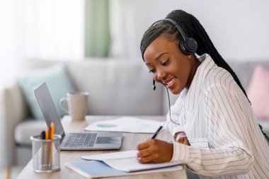 Evinden internet kursuna katılan neşeli Afro-Amerikalı genç kadın, dizüstü bilgisayar kullanıyor, not alıyor, Webinar dinliyor, yan görüş, kopyalama alanı dinliyor. Siyahi kız öğrenci online ders alıyor.