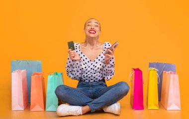 Nakit Kavramı. Kredi kartı ve akıllı telefon tutan, internetten sipariş veren, kıyafet alan, alışveriş torbalarıyla stüdyoda oturan heyecanlı stil sahibi bir kadın. Perakende ve Çevrimiçi Mağaza