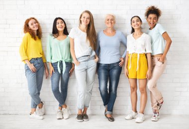 Kadın çeşitliliği mi? Beyaz tuğla duvarın arkasında duran kameraya gülümseyen çok ırklı kadınlar. Tam Uzunluk