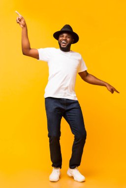 Şapkalı neşeli siyah adam dans ediyor ve sarı stüdyo arka planında işaret parmağıyla işaret ediyor.