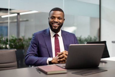 Modern ofisteki çalışma masasında poz veren takım elbiseli neşeli, yakışıklı genç siyahi iş adamı, dizüstü bilgisayarın önünde oturan Afro-Amerikan girişimci, kameraya gülümseyen, uzay kopyası